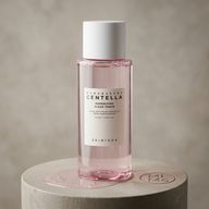 بطری شفاف تونر صورتی رنگ از برند اسکین ۱۰۰۴ با برچسب 'Madagascar Centella Poremizing Clear Toner' روی پایه بتنی قرار گرفته و مقداری تونر ریخته شده است.